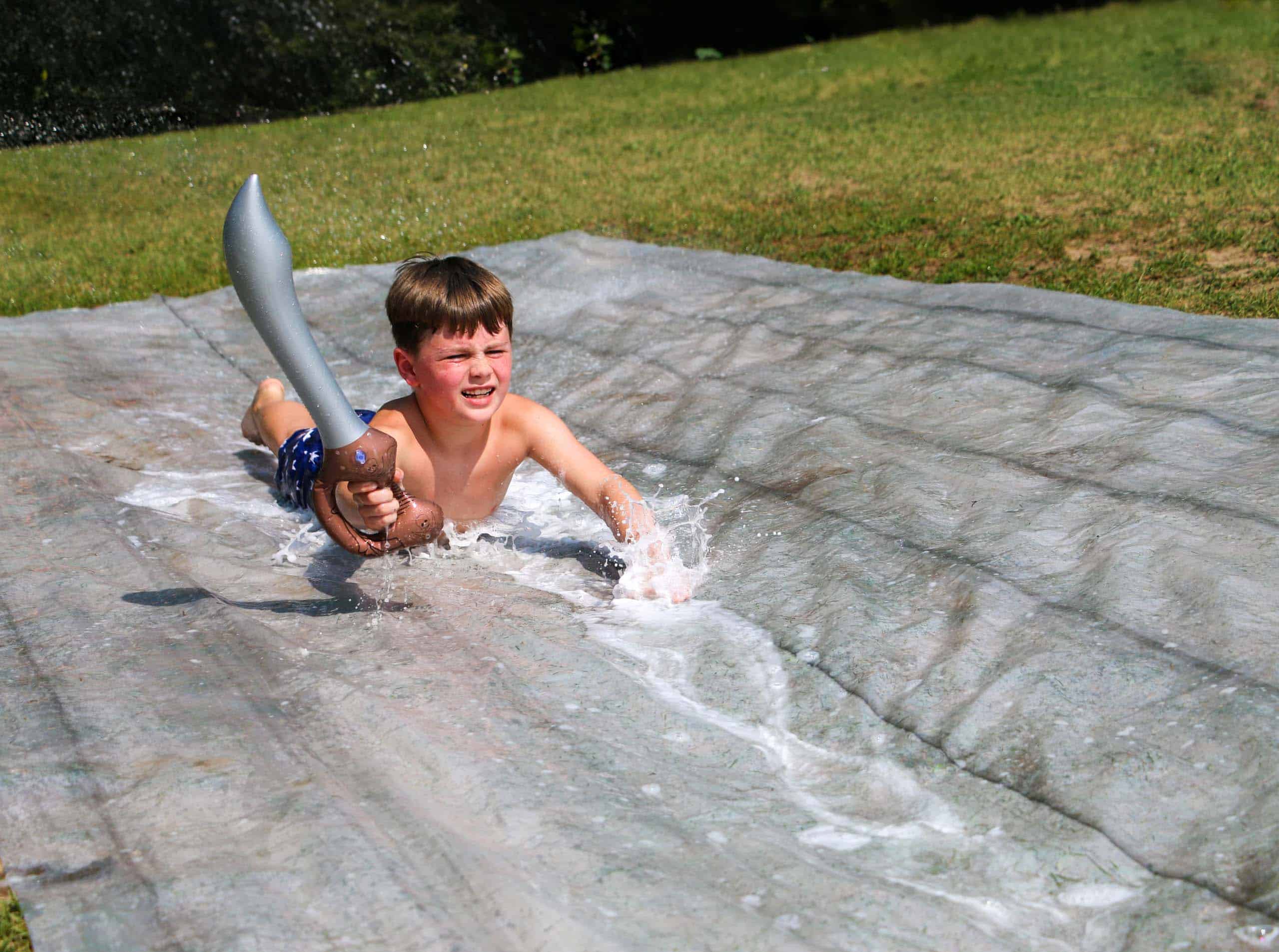 A boy sliding down a water slide at a summer camp in Iowa, holding a toy sword.