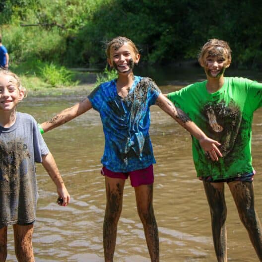 Three kids at a summer camp in Iowa standing in a river with mud on their faces.