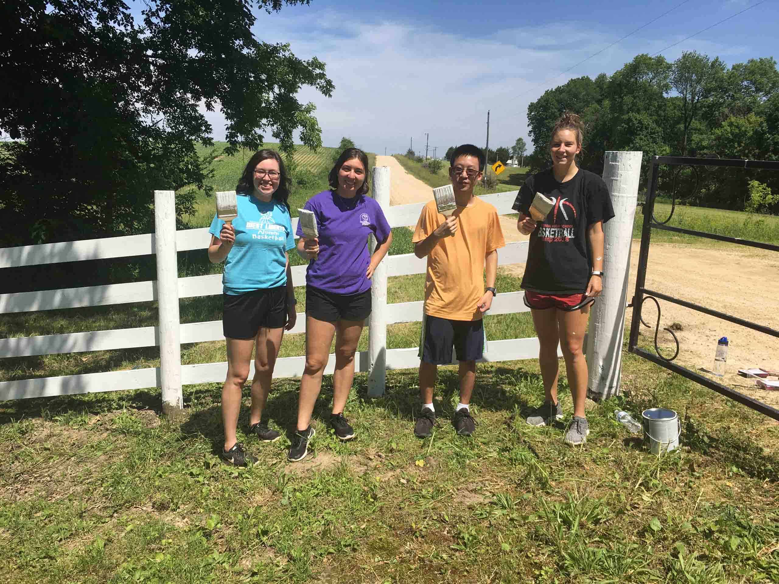 Four people standing with paintbrushes in front of a white fence at summer camp in Iowa.